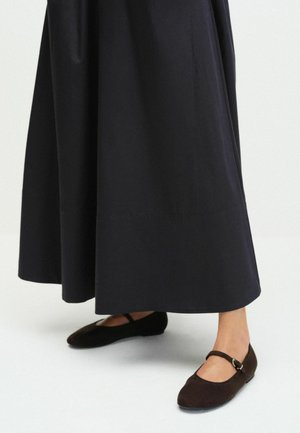 Person wearing black wide-leg pants and dark brown Mary Jane flat shoes standing on a light floor.