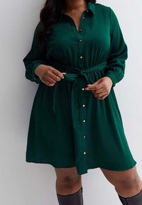 Green button-up dress made of smooth fabric, featuring a tied waist and long sleeves with button cuffs, complemented by gold-tone buttons.