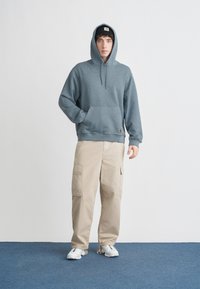 Bluza z kapturem w stonowanym niebieskim kolorze z prążkowaną teksturą, kieszenią z przodu i naszywką z logo. Połączona z beżowymi spodniami cargo i białymi trampkami.