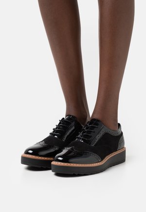 Gambe che indossano scarpe stringate in pelle verniciata nera e camoscio con suole spesse marroni e nere su uno sfondo bianco.