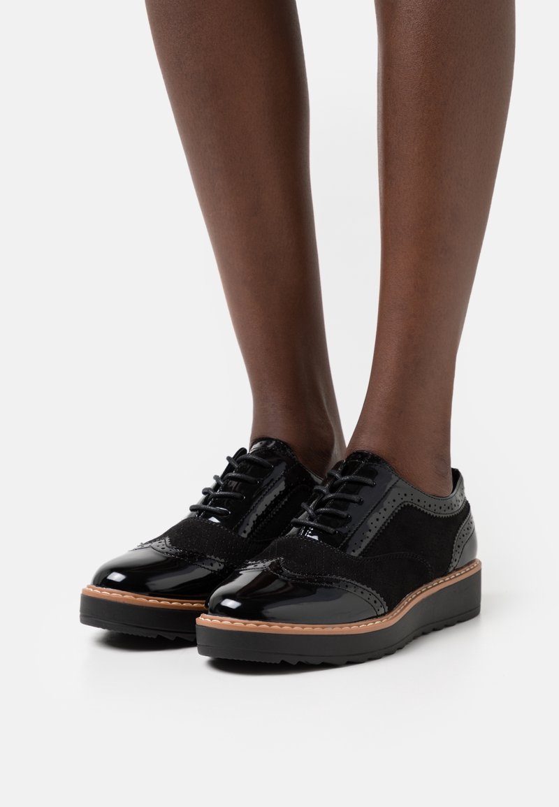 Ben som bär svarta lackläder- och mocka-wingtip-schnörskor med tjocka bruna och svarta sulor på vit bakgrund.