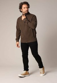 Veste zippée marron avec un col montant, des poches avant et des poignets côtelés, associée à un jean skinny noir et des baskets beiges avec des accents blancs.
