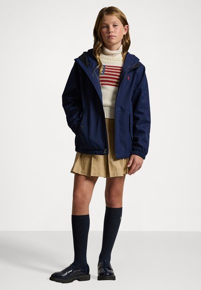 Marineblaue wasserdichte Jacke mit Kapuze, ausgestattet mit elastischen Bündchen, kombiniert mit einem cremefarbenen gestreiften Pullover, einem khakifarbenen Rock, marineblauen knielangen Socken und glänzenden Schuhen.