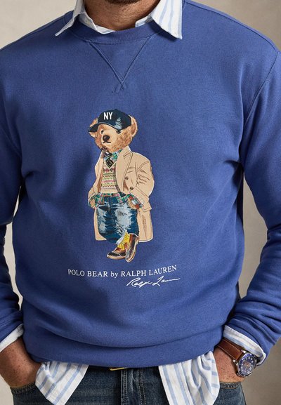 Άνδρας που φοράει μπλε φούτερ με γραφικό του Polo Bear ντυμένος με στρώσεις ρούχων και καπέλο NY, συνδυασμένο με ριγέ πουκάμισο και ρολόι χειρός.