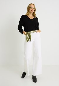 Top negro de manga larga con escote en v, vaqueros blancos de pierna ancha con dobladillo deshilachado, bufanda de seda verde con estampado anudada a la cintura, botines negros con tacón.