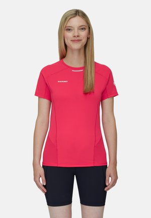 Kvinde står og smiler iført en lyserød kortærmet sportstrøje med "Mammut"-logo og sorte shorts mod en ensartet baggrund.