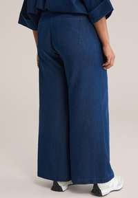 Wide-leg jeans iz temno modre barve, ki imajo dve zadnji žep in sproščen kroj. Material ima gladko teksturo, dopolnjuje ga podvrtan rob.