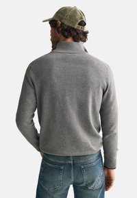 Grauer Strickpullover mit hohem Kragen, kombiniert mit blauen Jeans. Der Stoff wirkt weich, und das Design zeichnet sich durch eine taillierte Silhouette aus.