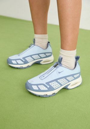 Osoba nosząca jasnoniebieskie i szare buty sportowe Nike z białymi skarpetami za kostkę stoi na zielonej podłodze.