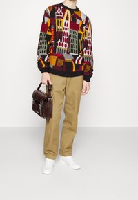 Suéter de lana colorido con patrones geométricos, combinado con pantalones beige y zapatillas blancas. Sostiene un bolso de cuero marrón.