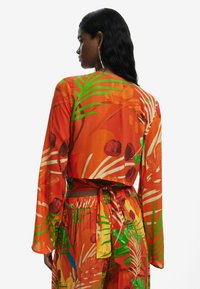 Blomsterindpakket top i livlig orange med grønne tropemønstre. Lavet af glat stof; har lange ærmer og et bindebånd bagpå.