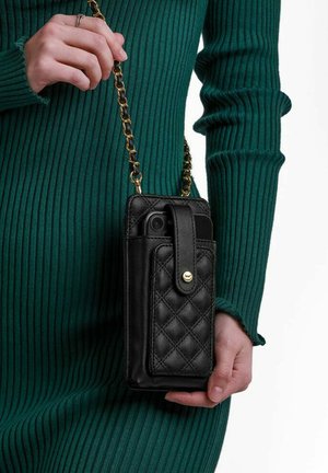 Schwarze gesteppte Lederhandytasche mit abnehmbarem Kettenriemen. Mit einer Fronttasche und einem Knopfverschluss. In der Hand gehalten gegen einen grünen gerippten Pullover.