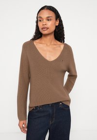 Rudas megztinis su V formos iškirpte, ilgomis rankovėmis ir prigludusia silueto forma. Maža logotipo detalė ant krūtinės. Derinama su tamsiai mėlynomis džinsomis.