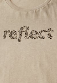 Camiseta beige con cuello redondo que presenta la palabra "reflect" adornada con tachuelas circulares negras, creando un diseño texturizado.