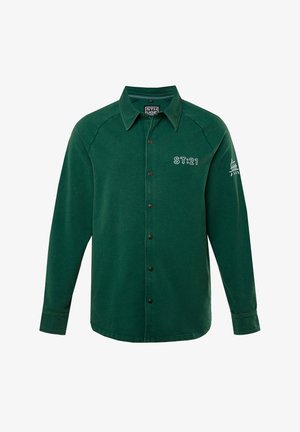 Zelená košile s dlouhým rukávem a knoflíkovým zapínáním, klasického límce, s akcenty na bočních švech a vyšitým detailem "ST:21" na levé hrudi a rukávu.
