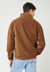 Chaqueta de pana marrón con un alto cuello, puños elásticos y un corte relajado. Superficie texturizada con estrías verticales. Superpuesta sobre una camisa roja.