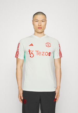 Weißes Fußballtrikot von Manchester United mit roten Akzenten, ausgestattet mit einem V-Ausschnitt, kurzen Ärmeln und den Logos von Tezos und Adidas.