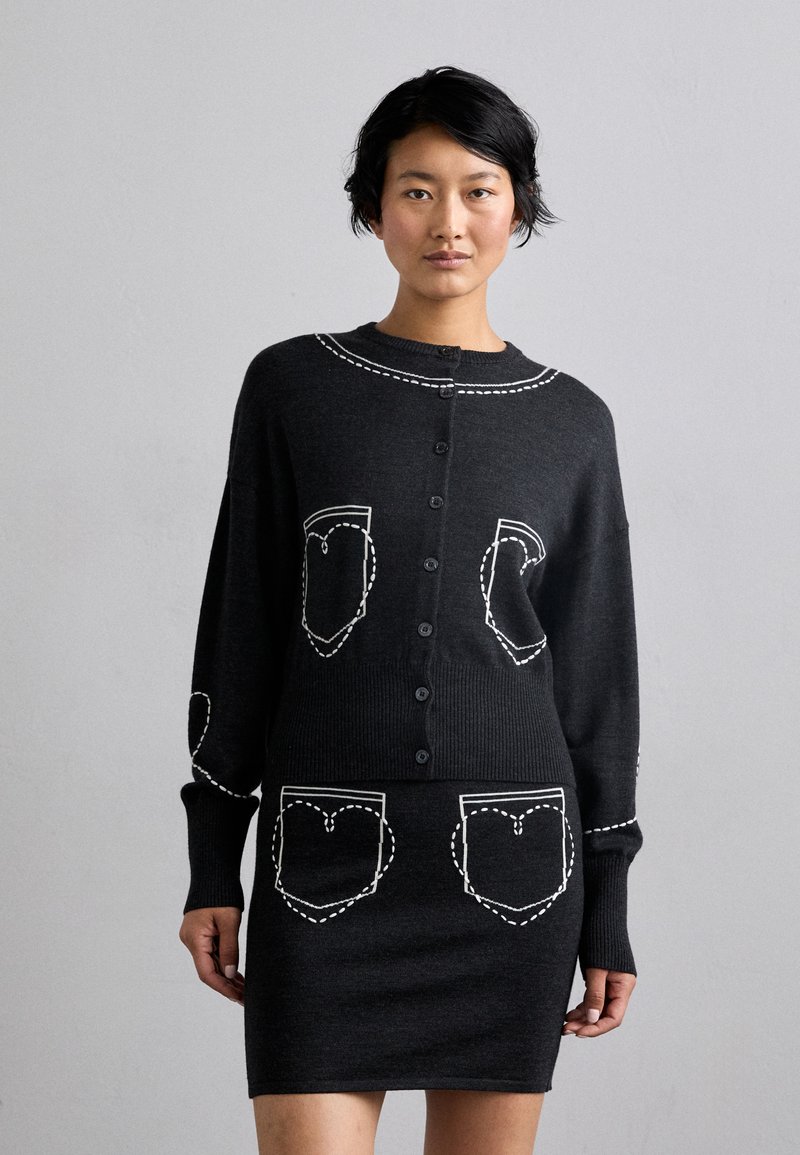 Fekete kötött kardigán fehér hímzett szív alakú zsebekkel, kerek nyakkivágással és gombos záródással. Puha tapintású, laza fazonú.