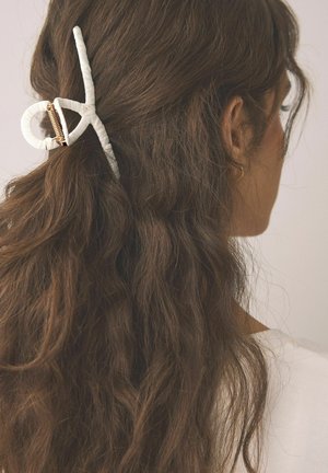 Weißer Haarklammer mit rundem Design, strukturierter Oberfläche und goldfarbenen Beschlägen, hält welliges braunes Haar zurück. Der Hintergrund ist neutral.