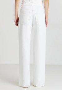 Persona che indossa pantaloni bianchi larghi, mostrata di spalle, in piedi contro uno sfondo grigio chiaro uniforme.