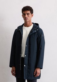 Jonge man in een donkerblauwe hoodie over een wit shirt, staand tegen een vlakke lichtgrijze muur.