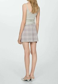 Jeune femme portant un débardeur gris, une mini-jupe trapèze à carreaux et des escarpins blancs à petit talon, debout devant un fond clair uni.