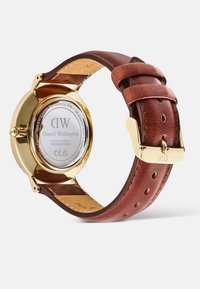 Reloj con correa de cuero marrón y caja de acero inoxidable dorado, forma redonda y esfera blanca con la marca 'DW'. Incluye cierre de hebilla.