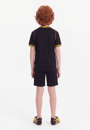 Enfant aux cheveux roux bouclés, de dos, portant une tenue de sport noire avec des bordures jaunes et des chaussures de football noires et jaunes.