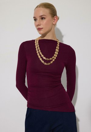 Kvinde med blondt hår sat op i en knold iført en tætsiddende burgunderfarvet langærmet top, marineblå bukser og en lagdelt guldkæde, stående foran en hvid væg.