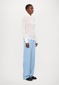Langärmliges, enges weißes Oberteil mit einem Wasserfallausschnitt, kombiniert mit weitem hellblauen Hosen. Das Modell trägt schwarze Schuhe und hat kurze Haare.