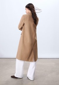 Langer beige Wollmantel mit geradem Schnitt und einem Schlitz hinten. Kombiniert mit weiten weißen Hosen und dunklen Knöchelstiefeln. Sauberes, minimalistisches Design.