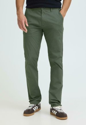 Mann trägt olivgrüne Straight-Fit-Hose mit Seitentaschen und schwarze Turnschuhe mit weißen Streifen, steht vor weißem Hintergrund.