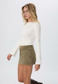 Top a maniche lunghe bianco, aderente e con texture a coste, abbinato a una mini gonna di jeans verde oliva con tasche frontali e chiusura a bottone.