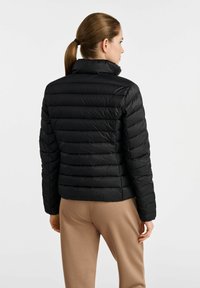 Czarna kurtka puchowa z pikowanym wzorem, wysokim kołnierzem i dopasowanym krojem. Noszona z beżowymi spodniami, eksponując lekką, gładką fakturę.