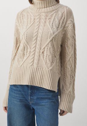 Femme portant un pull à col roulé beige en maille torsadée avec des poignets et un ourlet côtelés, associé à un jean bleu.