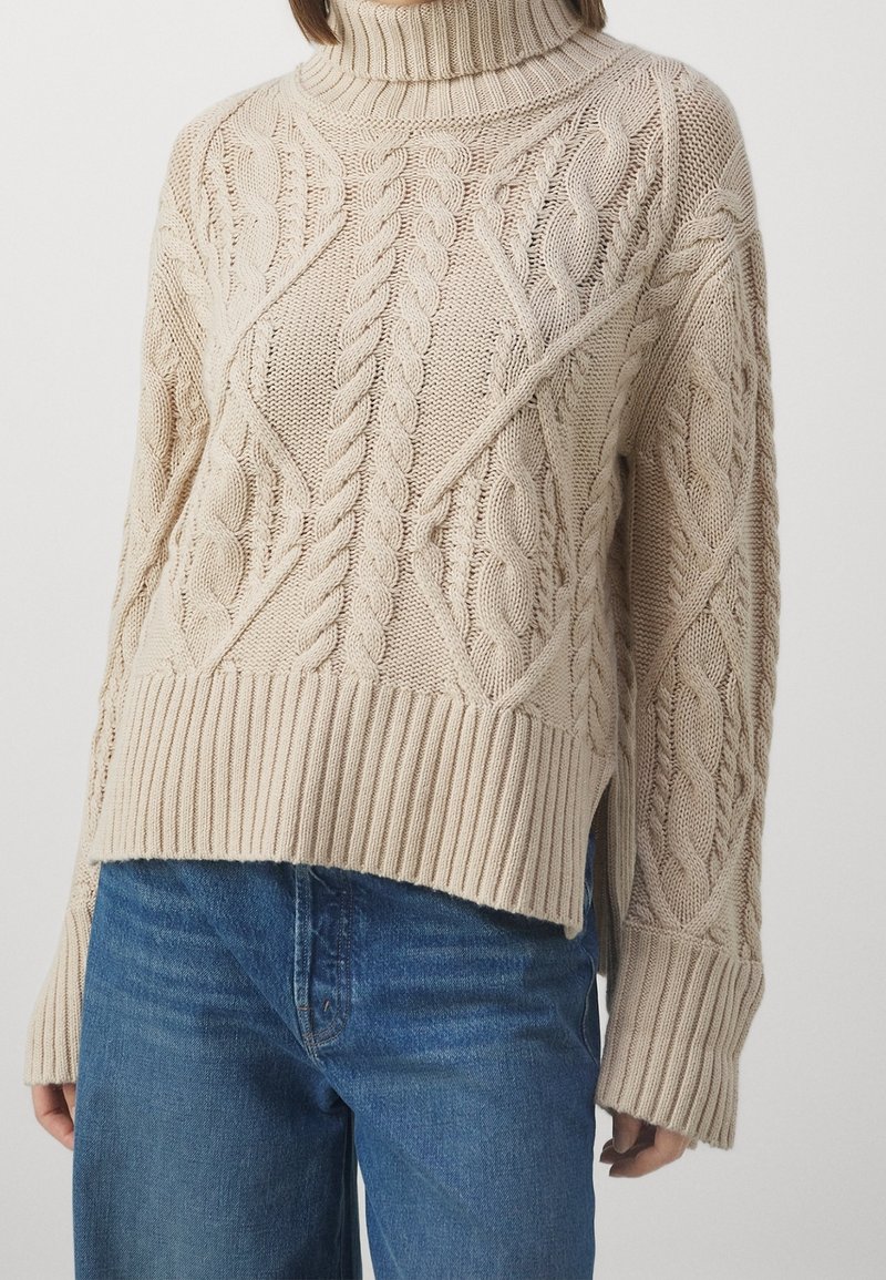 Femme portant un pull à col roulé beige en maille torsadée avec des poignets et un ourlet côtelés, associé à un jean bleu.