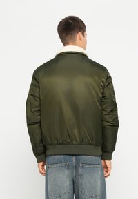 Veste bomber rembourrée de couleur vert olive avec un col en fausse fourrure blanche, des poignets et une taille côtelés ; présente un extérieur lisse et brillant.