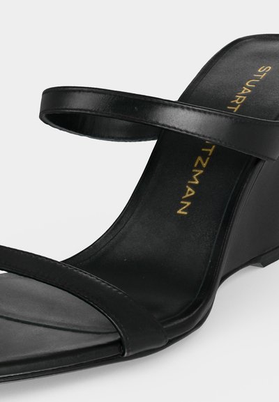Sandale compensée en cuir noir avec deux fines brides et marquage doré "Stuart Weitzman" sur la semelle intérieure, présentée sur fond blanc.