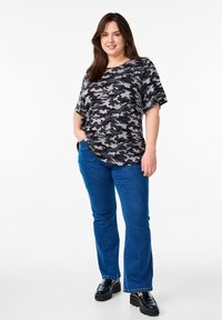 T-shirt à motifs camouflage noir et gris avec des manches courtes, associé à un jean bleu et des chaussures à plateforme noires. Le mannequin se tient avec une main dans la poche.