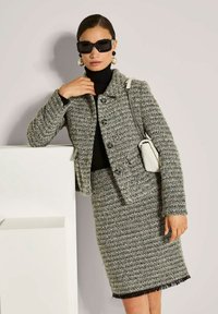 Frau in grauem Tweedjackett und passendem Rock, schwarzem Rollkragenpullover, übergroßen schwarzen Sonnenbrillen und Perlenohrringen, die eine weiße gesteppte Tasche hält.