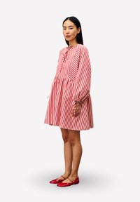 Eine Frau steht in einem rot-weiß gestreiften Kleid mit langen Ärmeln und roten Mary Jane Schuhen vor einem einfarbigen weißen Hintergrund.