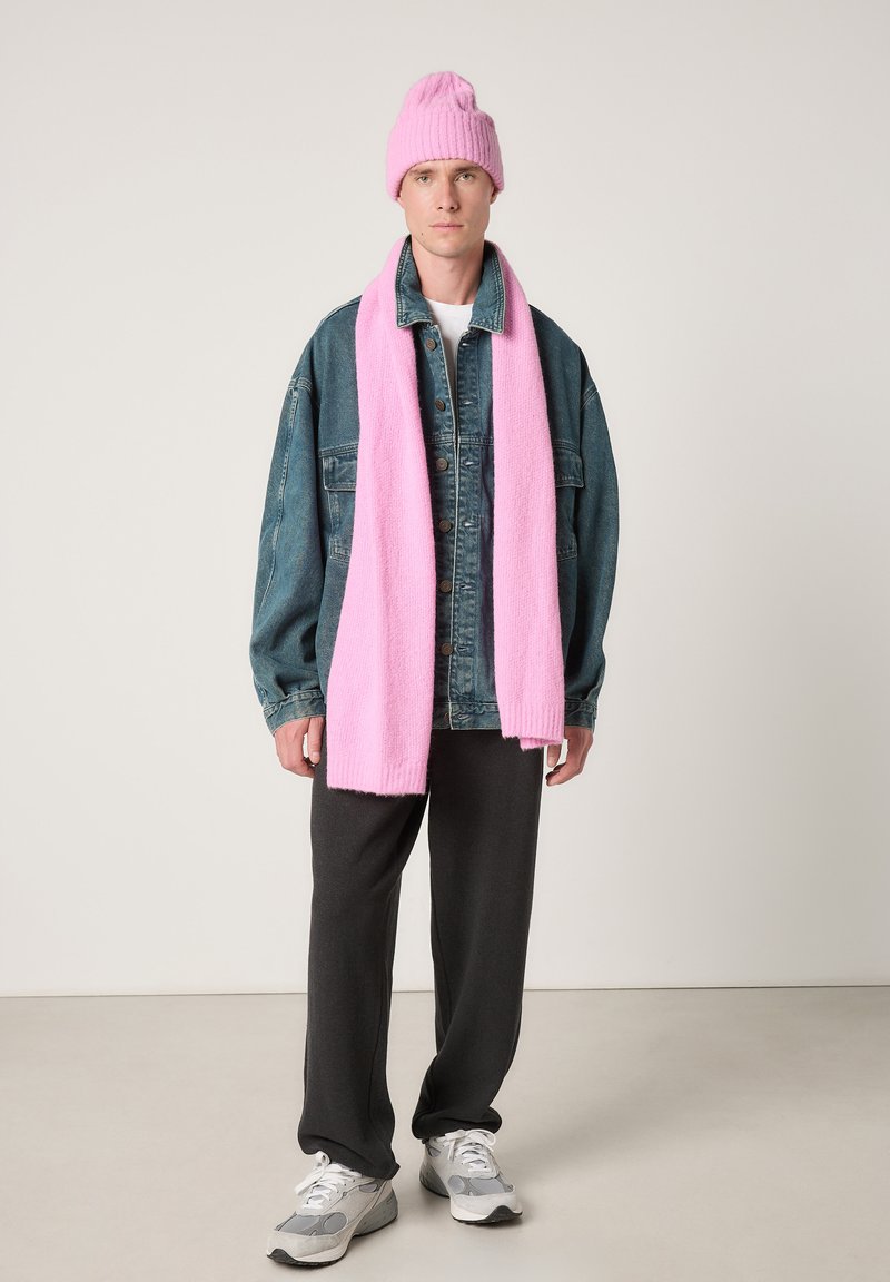 Spijkerjack, wit shirt, grijze joggingbroek, grijze sneakers, roze gebreide muts en roze gebreide sjaal op een neutrale achtergrond.