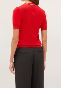 Rød t-shirt med korte ærmer og tekstureret overflade, prydet med en dekorativ krave med perlekant og en nøglehulslukning i ryggen. Matchet med sorte bukser.