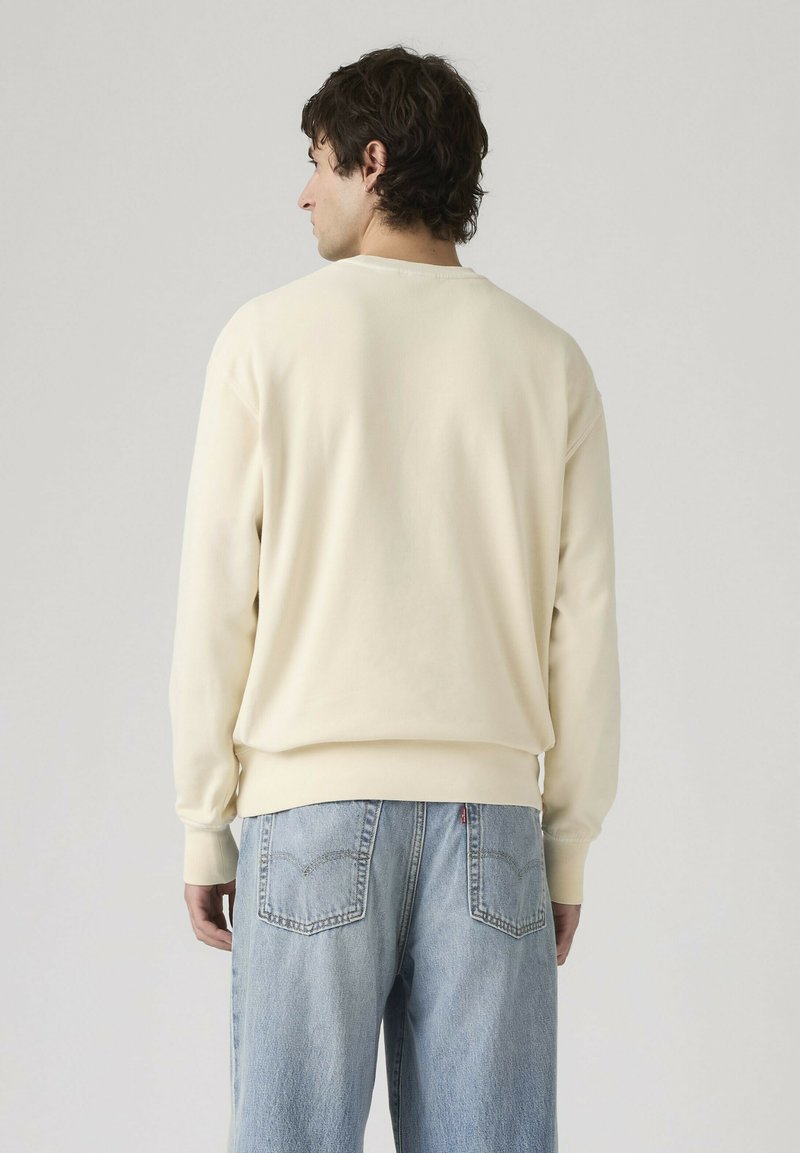 Uomo con capelli scuri che indossa una felpa color crema e jeans azzurri, visto di spalle su uno sfondo semplice grigio chiaro.