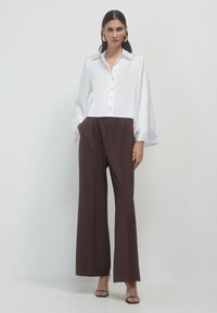 Donna in piedi che indossa una camicia bianca con bottoni, pantaloni larghi neri, sandali neri e grandi orecchini ad anello su sfondo neutro.