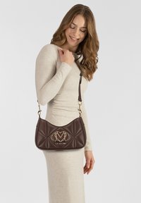Donna in abito beige porta una borsa a tracolla trapuntata marrone scuro con logo dorato e tracolla regolabile su sfondo neutro.