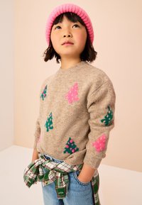 Maglione lavorato a maglia beige con motivi di alberi verdi e rosa. Abbinato a una camicia di flanella a quadri annodata in vita e jeans di denim blu.