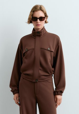 Vrouw draagt een bruin bijpassend jasje en broek met knopdetails en zwarte rechthoekige zonnebril, staat tegen een effen lichte muur.