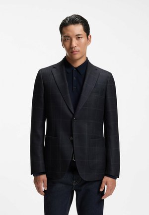 Mann trägt einen dunkel karierten Blazer über einem schwarzen Hemd mit Knöpfen und dunklen Jeans, steht vor einem schlichten weißen Hintergrund.