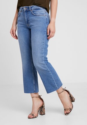 Frau trägt hellblaue, kurze Jeans und braune High-Heeled-Sandalen mit Schlangenmuster, steht auf weißem Boden mit neutralem Hintergrund.